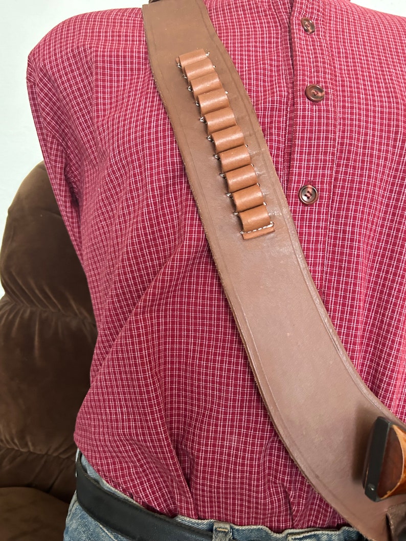 Pode incluir: Um coldre de pistola de couro marrom com um la&ccedil;o de muni&ccedil;&atilde;o de couro marrom. O coldre &eacute; usado sobre uma camisa xadrez vermelha e branca.