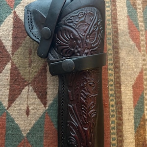 May include: A black leather holster with intricate floral tooling. The holster features a belt loop and a strap with a snap closure. The leather has a rich, dark color and a detailed embossed design.