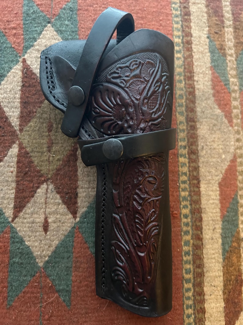May include: A black leather holster with intricate floral tooling. The holster features a belt loop and a strap with a snap closure. The leather has a rich, dark color and a detailed embossed design.