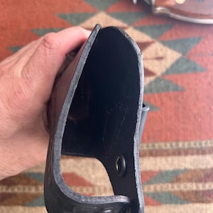 May include: A black leather holster with a riveted design. The holster is open at the top and has a loop for a belt. A silver revolver with a brown handle is visible in the background.