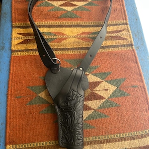 May include: Black leather shoulder holster with a decorative floral pattern. The holster has a strap that goes over the shoulder and around the back. The holster is designed to hold a handgun.