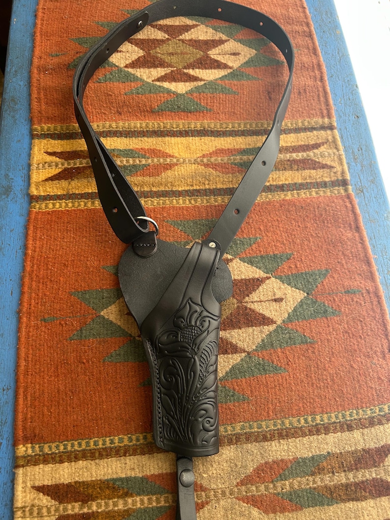 May include: Black leather shoulder holster with a decorative floral pattern. The holster has a strap that goes over the shoulder and around the back. The holster is designed to hold a handgun.