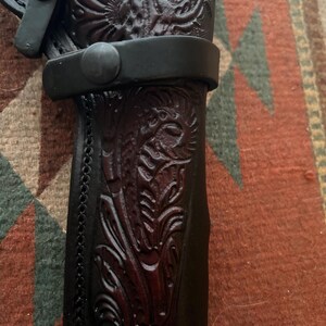 May include: Close-up of a black leather holster with intricate floral and animal carvings. The holster features a stitched edge and a black strap with a button closure. The leather has a dark brown hue in the carved areas.