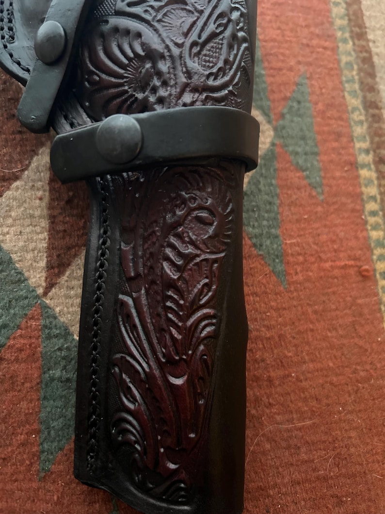 May include: Close-up of a black leather holster with intricate floral and animal carvings. The holster features a stitched edge and a black strap with a button closure. The leather has a dark brown hue in the carved areas.