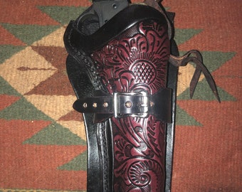 Peut inclure: Un étui en cuir noir avec un travail floral complexe. L'étui a une boucle de ceinture et une boucle. Un revolver noir et or est à l'intérieur de l'étui.