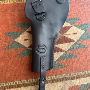 May include: Black leather holster with a shoulder strap. The holster has a stitched design and a stamped logo that reads "The Saddle Maker, Paso". The holster is designed to hold a firearm.