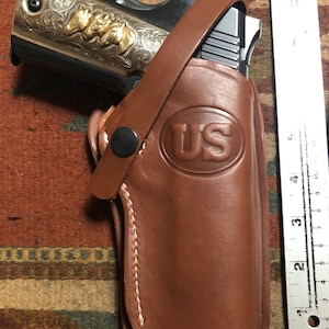 May include: A brown leather holster with a stitched edge and a US logo embossed on the front. A silver and gold handgun is partially visible inside the holster.
