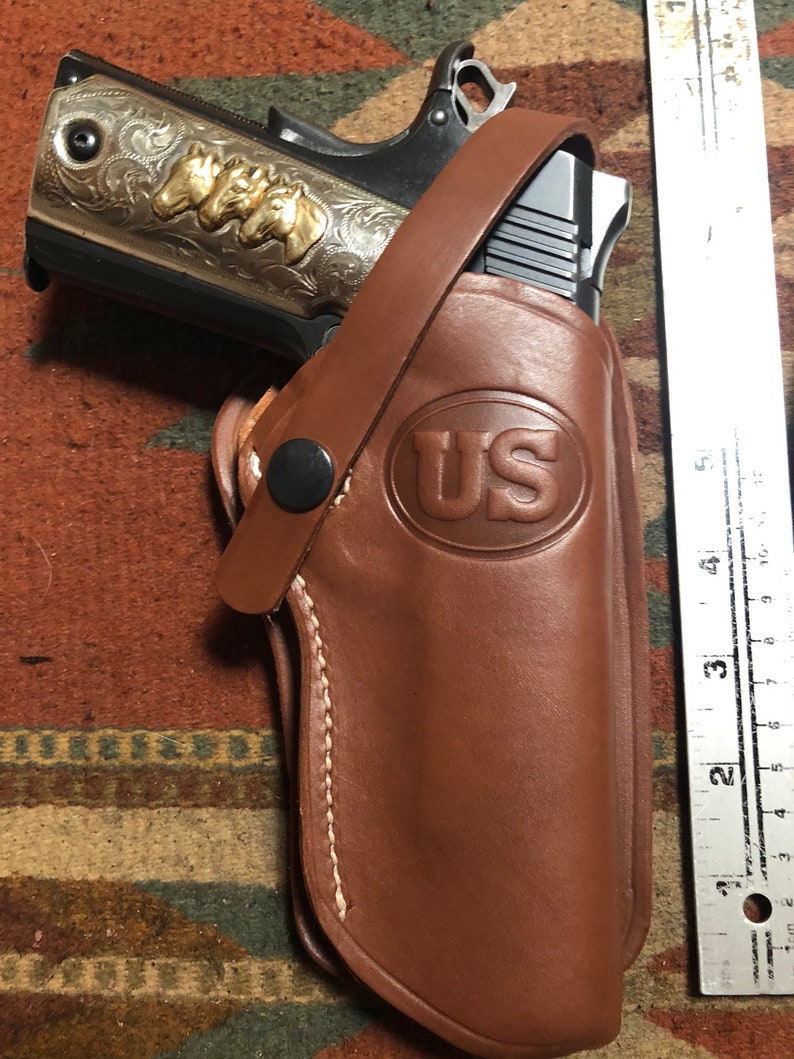 May include: A brown leather holster with a stitched edge and a US logo embossed on the front. A silver and gold handgun is partially visible inside the holster.