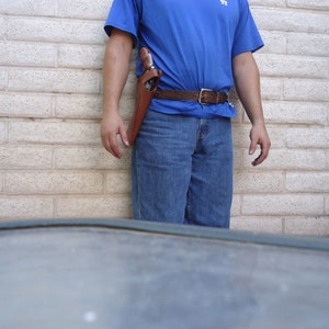 Puede incluir: Una persona con una camisa azul y pantalones vaqueros con una funda de cuero marr&oacute;n y una pistola sujeta a su cintur&oacute;n.