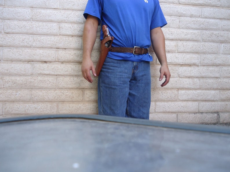 Puede incluir: Una persona con una camisa azul y pantalones vaqueros con una funda de cuero marr&oacute;n y una pistola sujeta a su cintur&oacute;n.