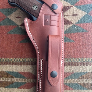 May include: A brown leather holster containing a black semi-automatic pistol with a textured grip and a Ruger logo. The holster features a stamped Texas flag design, a retention strap with a black snap button, and an integrated magazine pouch.