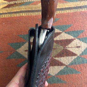 May include: A black leather holster with a basketweave pattern, holding a revolver with a brown wooden grip. The holster is open, revealing the silver barrel of the firearm.