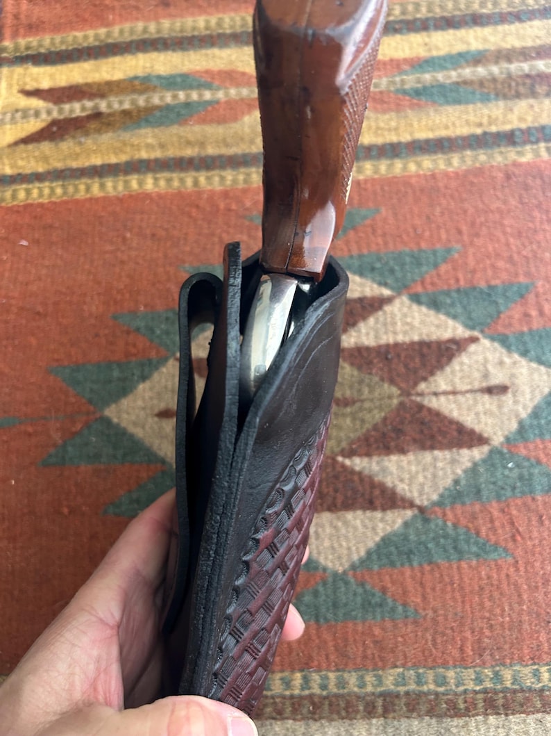 May include: A black leather holster with a basketweave pattern, holding a revolver with a brown wooden grip. The holster is open, revealing the silver barrel of the firearm.