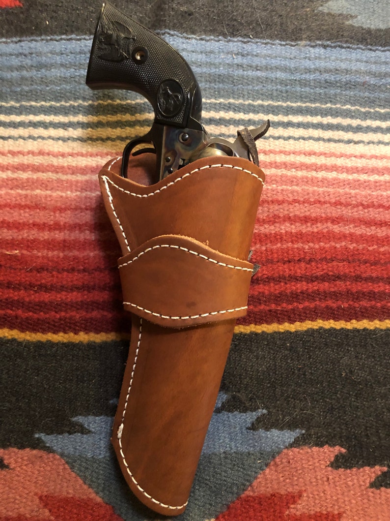 May include: A brown leather holster with white stitching, holding a black revolver with a silver barrel. The holster is on a colorful striped blanket.