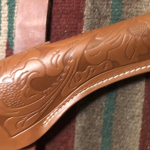 May include: A brown leather holster with a decorative floral design. The holster has a white stitching detail around the edge.