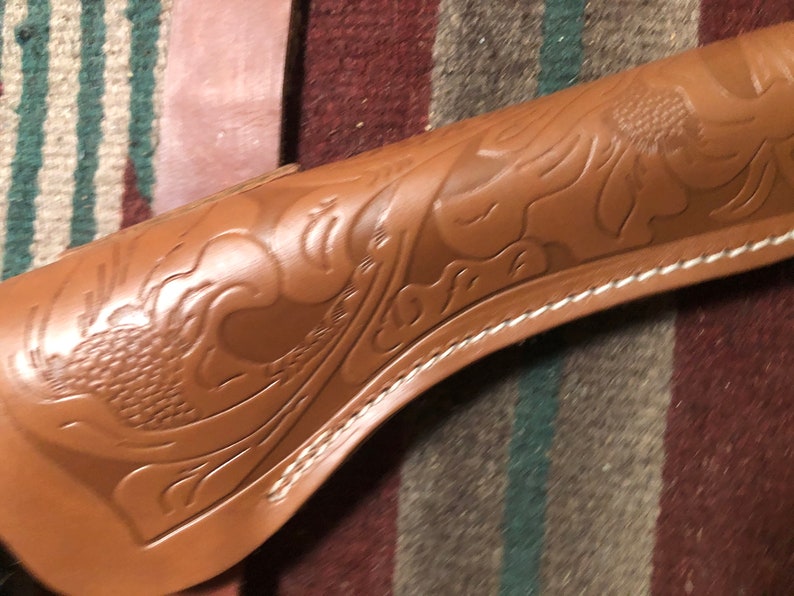 May include: A brown leather holster with a decorative floral design. The holster has a white stitching detail around the edge.