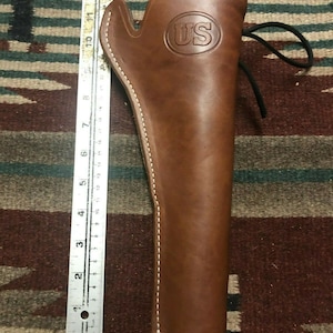 Op de afbeelding: Een bruine leren holster met witte stiksels en een US-logo. De holster heeft een band met een gesp.