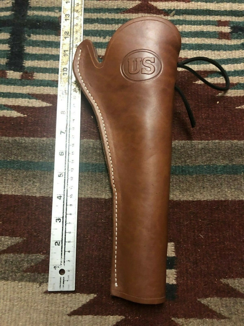 Op de afbeelding: Een bruine leren holster met witte stiksels en een US-logo. De holster heeft een band met een gesp.