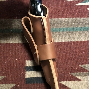 Op de afbeelding: Een bruine leren holster met een zwart pistool erin. De holster heeft een band die om de taille gaat.