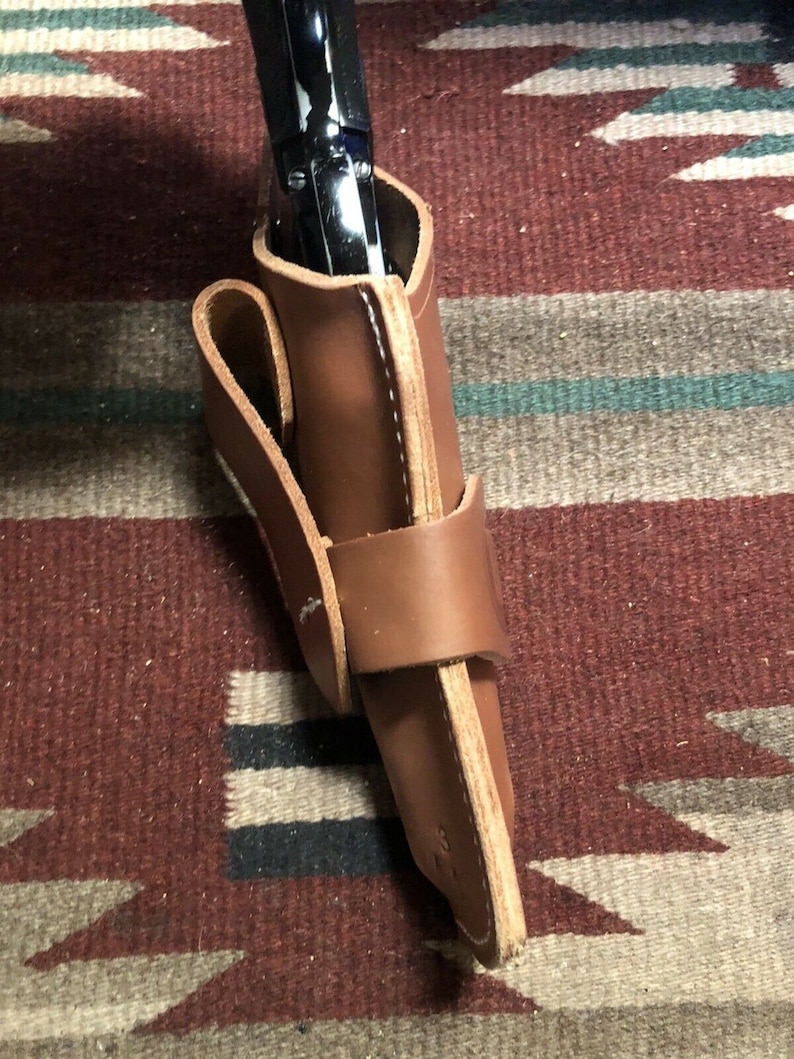 Op de afbeelding: Een bruine leren holster met een zwart pistool erin. De holster heeft een band die om de taille gaat.