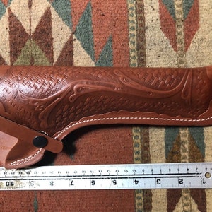 Op de afbeelding: Een bruine leren holster met een geweven patroon en witte stiksels. De holster is 23 cm lang.