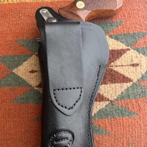 May include: A black leather holster with a stitched design, holding a revolver with a brown wooden grip. The holster has a stamped logo that reads "SADDLE MAKER". The revolver's grip has a gold-coloured medallion.
