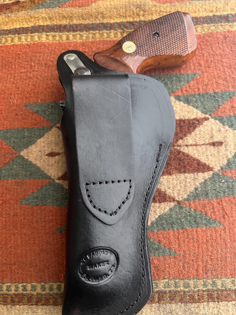 May include: A black leather holster with a stitched design, holding a revolver with a brown wooden grip. The holster has a stamped logo that reads "SADDLE MAKER". The revolver's grip has a gold-coloured medallion.