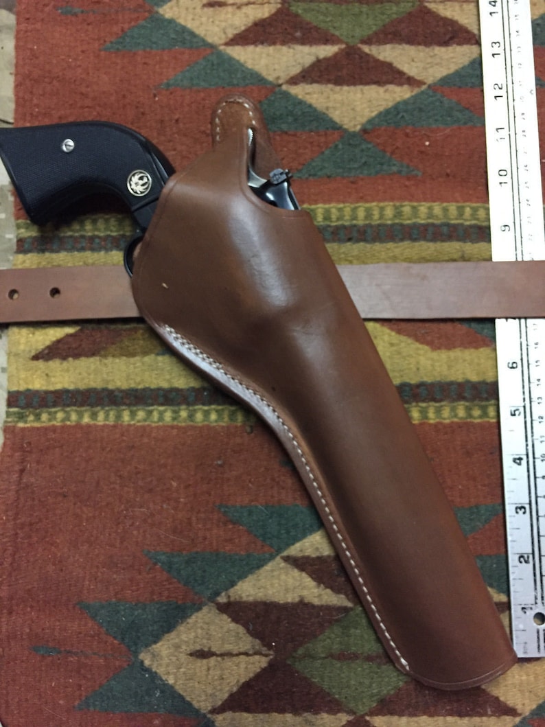Puede incluir: Una funda de cuero marr&oacute;n con una pistola negra en su interior. La funda tiene un detalle de costura blanca y est&aacute; sobre una alfombra estampada.
