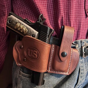 Könnte beinhalten: Ein braunes Lederholster mit eingeprägtem US-Logo und integrierter Magazintasche, das eine schwarze halbautomatische Pistole mit verzierten goldfarbenen Griffschalen mit Pferdekopf-Gravuren enthält, getragen am Gürtel über Jeans und einem rot karierten Hemd.