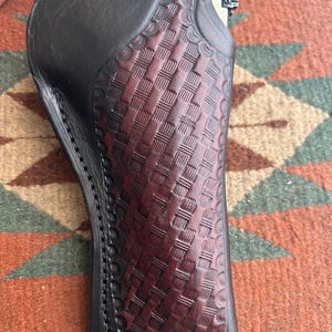 May include: A black and burgundy leather holster with a basketweave pattern. The holster has a scalloped edge and is stitched with black thread. The leather is a deep burgundy colour in the centre and black on the edges.