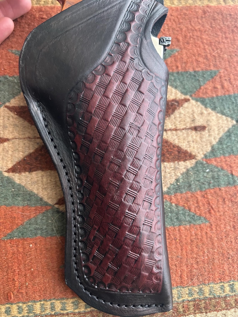 May include: A black and burgundy leather holster with a basketweave pattern. The holster has a scalloped edge and is stitched with black thread. The leather is a deep burgundy colour in the centre and black on the edges.