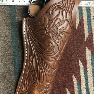 Op de afbeelding: Een bruine leren holster met ingewikkelde bloemmotieven. De holster heeft een witte stikdetail rond de randen. De holster is ontworpen om een ​​vuurwapen te houden.