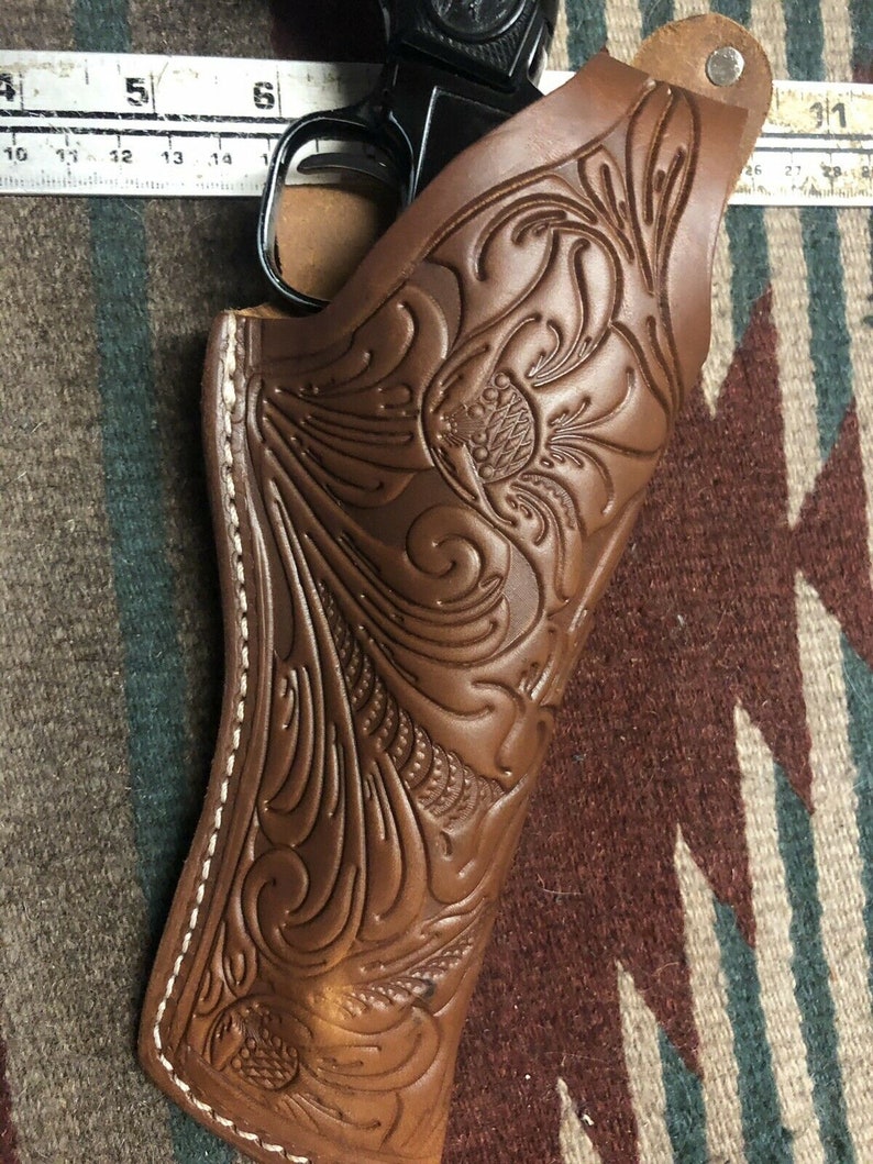 Op de afbeelding: Een bruine leren holster met ingewikkelde bloemmotieven. De holster heeft een witte stikdetail rond de randen. De holster is ontworpen om een ​​vuurwapen te houden.