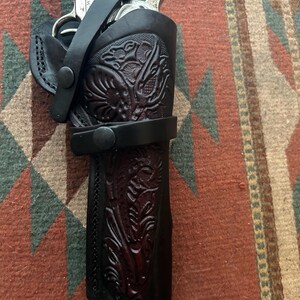 May include: A black leather holster with intricate floral tooling, holding a revolver. The holster features a strap with a snap closure and is set against a patterned rug.