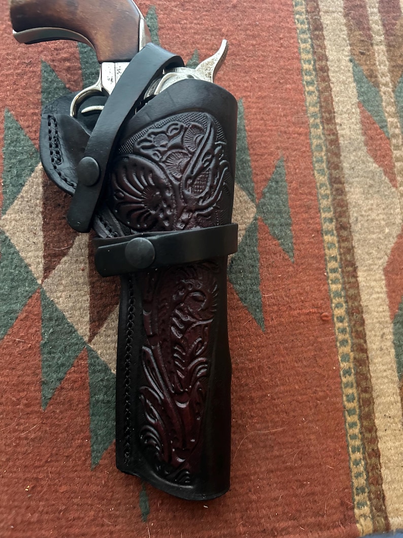 May include: A black leather holster with intricate floral tooling, holding a revolver. The holster features a strap with a snap closure and is set against a patterned rug.