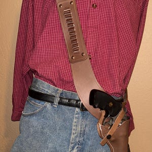 Può includere: Una fondina da spalla in pelle marrone con cintura abbinata e un revolver. La fondina ha un cinturino con anelli per proiettili. Il modello indossa una camicia a quadri rossa e jeans blu.