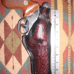 Könnte beinhalten: Ein braunes Lederholster mit aufwendigen Blumenmustern, entworfen, um einen silbernen Revolver zu halten. Das Holster ist 17,8 cm hoch und hat schwarze Nähte.