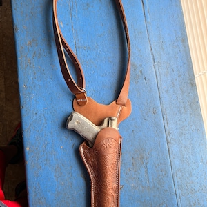 Puede incluir: Una funda de cuero marr&oacute;n con un intrincado dise&ntilde;o floral. La funda est&aacute; unida a una correa de hombro de cuero marr&oacute;n. Una pistola plateada est&aacute; dentro de la funda.