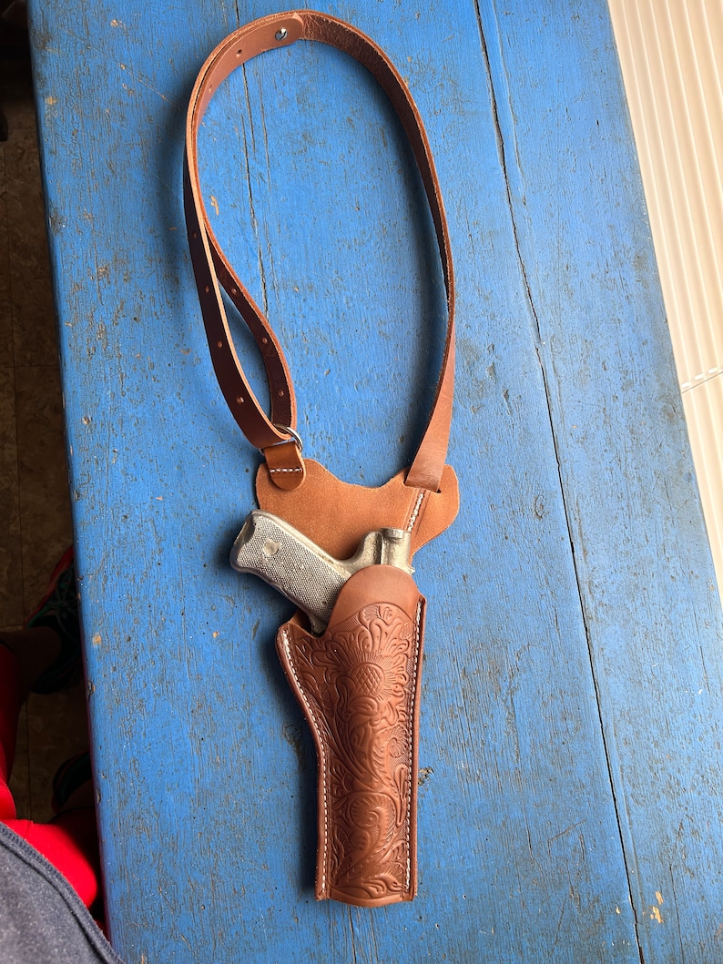 Puede incluir: Una funda de cuero marr&oacute;n con un intrincado dise&ntilde;o floral. La funda est&aacute; unida a una correa de hombro de cuero marr&oacute;n. Una pistola plateada est&aacute; dentro de la funda.
