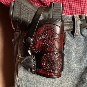 May include: A black handgun in a burgundy leather holster with floral and circular embossed designs. The holster is attached to a black belt worn with blue jeans. The red shirt is visible in the background.