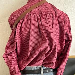 Puede incluir: Una camisa de manga larga a cuadros rojos y blancos con una correa de cuero marr&oacute;n en la espalda.