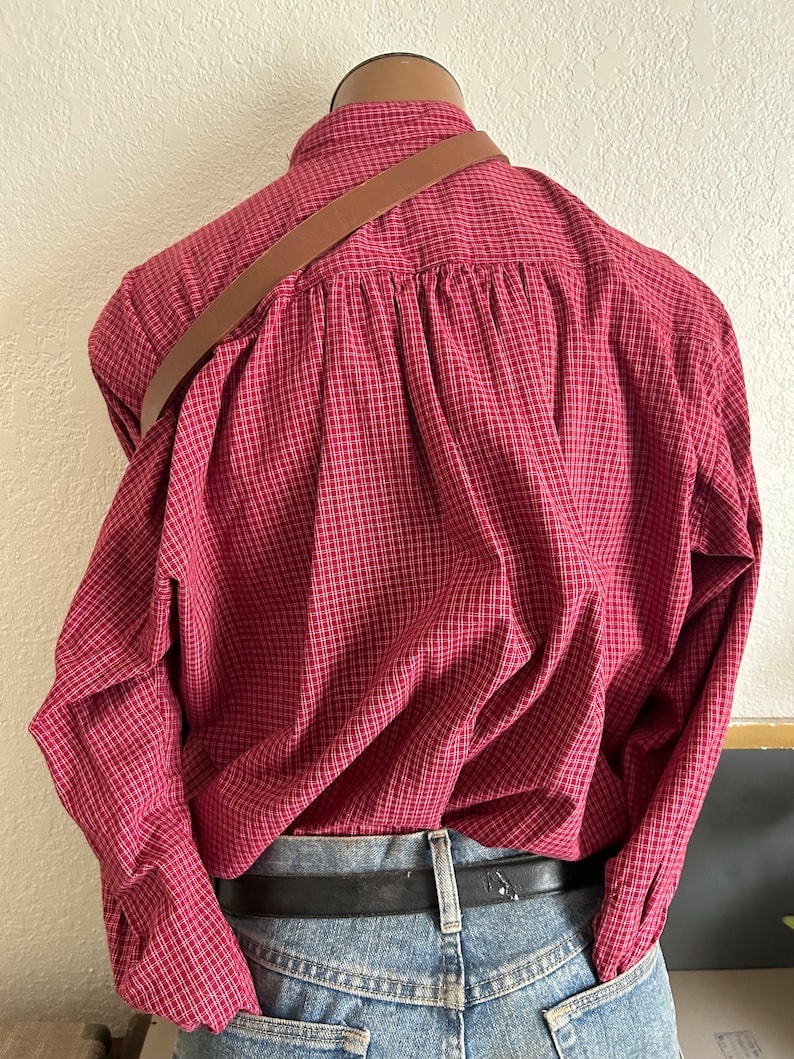 Puede incluir: Una camisa de manga larga a cuadros rojos y blancos con una correa de cuero marr&oacute;n en la espalda.