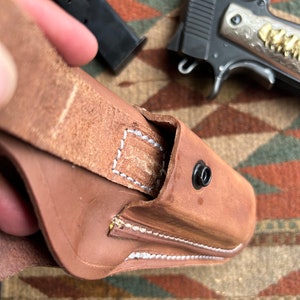May include: A brown leather holster with white stitching detail and a snap closure. The holster is designed to hold a handgun.