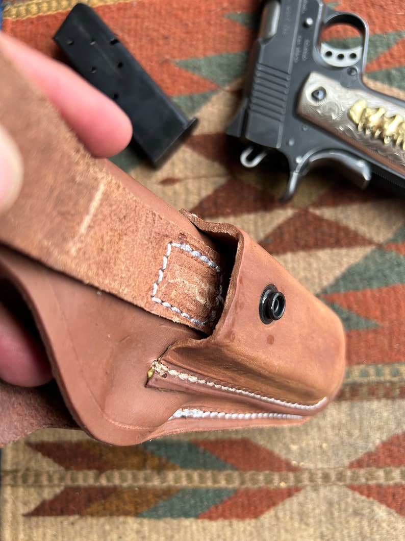 May include: A brown leather holster with white stitching detail and a snap closure. The holster is designed to hold a handgun.