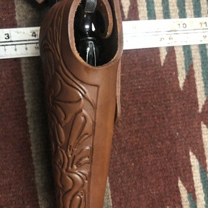 Op de afbeelding: Een bruine leren holster met ingewikkelde bloemmotieven. De holster is ontworpen om een ​​vuurwapen te dragen, met de loop van het wapen zichtbaar aan de bovenkant.