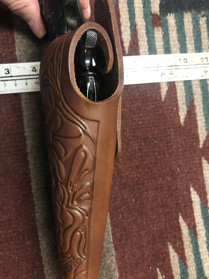 Op de afbeelding: Een bruine leren holster met ingewikkelde bloemmotieven. De holster is ontworpen om een ​​vuurwapen te dragen, met de loop van het wapen zichtbaar aan de bovenkant.