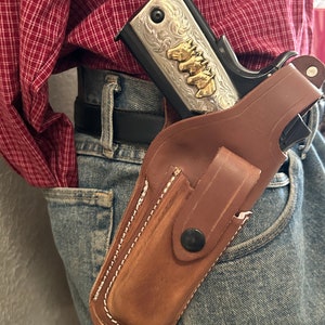 May include: A brown leather holster with a silver and gold engraved handgun inside. The holster is worn on a person's hip with a belt.