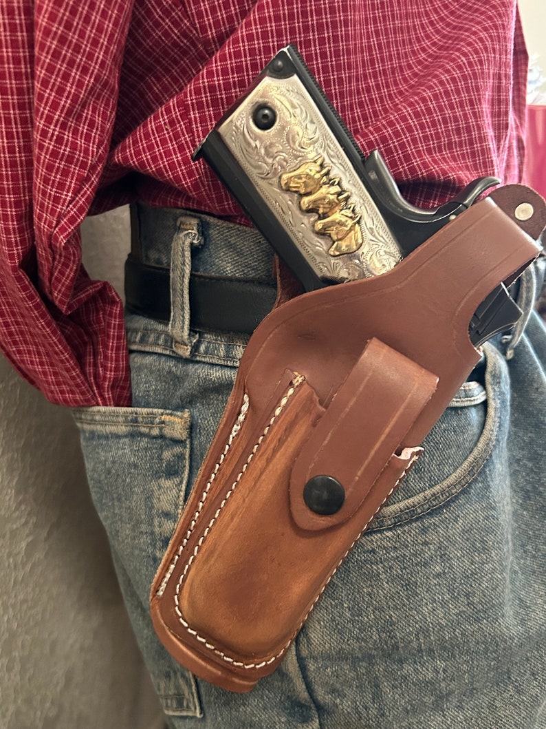 May include: A brown leather holster with a silver and gold engraved handgun inside. The holster is worn on a person's hip with a belt.