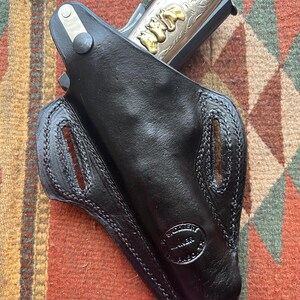 K&ouml;nnte beinhalten: Ein schwarzes Lederholster mit einer silber- und goldgravierten Pistole im Inneren. Das Holster hat ein gen&auml;htes Design und ein Herstellerzeichen mit der Aufschrift "WESTERN GUNNER".