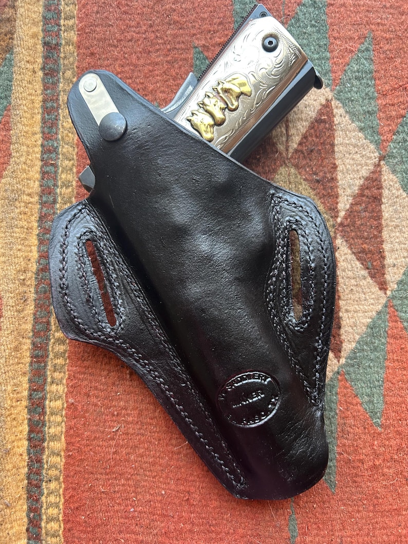 K&ouml;nnte beinhalten: Ein schwarzes Lederholster mit einer silber- und goldgravierten Pistole im Inneren. Das Holster hat ein gen&auml;htes Design und ein Herstellerzeichen mit der Aufschrift "WESTERN GUNNER".